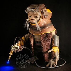 May include: A detailed prop of an alien-like creature with a brown and yellow helmet, fur-lined coat, and a cigarette. The creature holds a futuristic tool with a blue light. The prop is from Caliber Craft Props.
