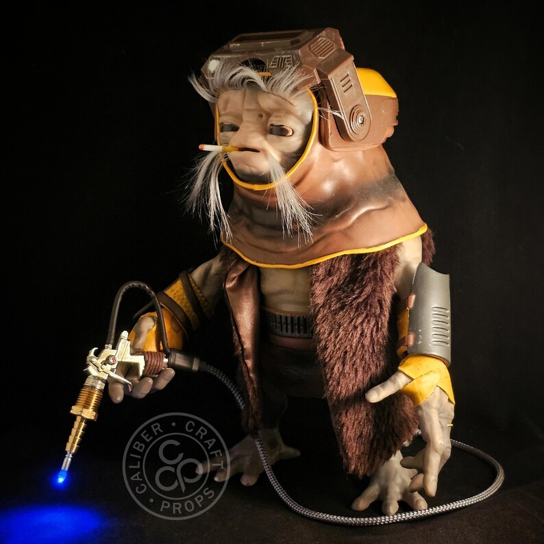 May include: A detailed prop of an alien-like creature with a brown and yellow helmet, fur-lined coat, and a cigarette. The creature holds a futuristic tool with a blue light. The prop is from Caliber Craft Props.