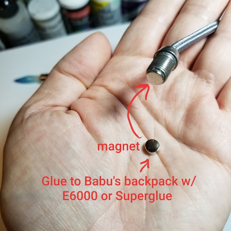 May include: A close-up of a small, round, black magnet. The magnet is being held in a hand. The magnet is attached to a silver metal rod. The text "Glue to Babu's backpack w/ E6000 or Superglue" is written below the magnet.