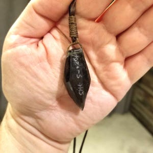 Puede incluir: Un colgante negro con un cordón de cuero marrón. El colgante tiene forma de óvalo puntiagudo y tiene escritura en él.