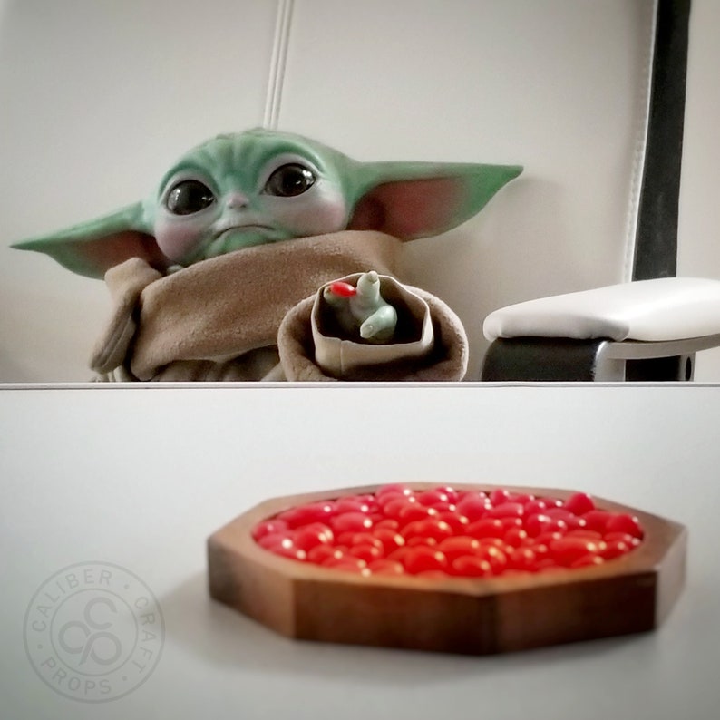 May include: A green-skinned alien character with large eyes and pointed ears, wrapped in a tan blanket, with a small toy in its hand. A wooden, octagonal bowl filled with red candies sits in front of the character.