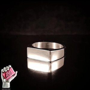 May include: A silver geometric ring with a rectangular design. The ring is highly reflective, with a mirrored reflection below. The background is black. A small, pink soap bar with the text "FIGHT CLUB" is held in a hand.