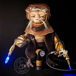 May include: A detailed prop figure of a character with a brown helmet, fur-lined coat, and yellow accents. The figure holds two futuristic tools with glowing blue lights. The Caliber Craft Props logo is visible.