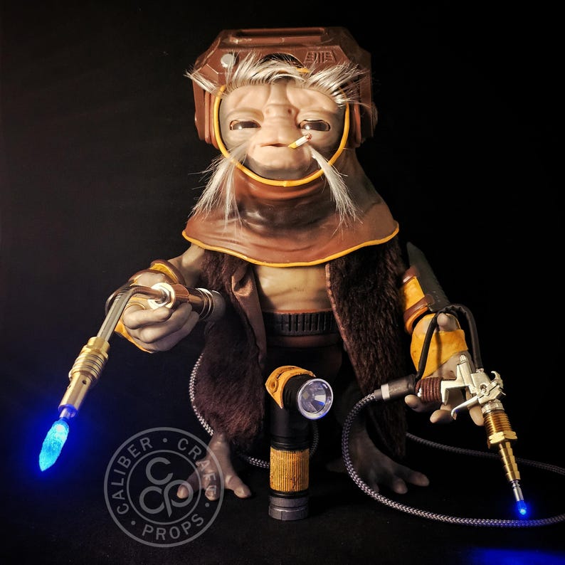May include: A detailed prop figure of a character with a brown helmet, fur-lined coat, and yellow accents. The figure holds two futuristic tools with glowing blue lights. The Caliber Craft Props logo is visible.