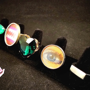 May include: A selection of rings on black velvet stands. The rings showcase diverse designs: a green stone, a geometric pattern, a heart-shaped emerald, an eye, and a silver geometric shape. The rings are presented against a dark backdrop.