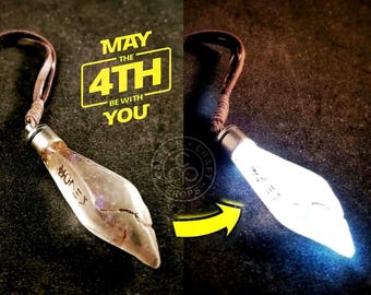 May include: A clear glass pendant with a brown leather cord. The pendant is shaped like a teardrop and has a silver cap on the top. The pendant glows white when lit. The text "May the 4th Be With You" is printed in yellow on a black background.