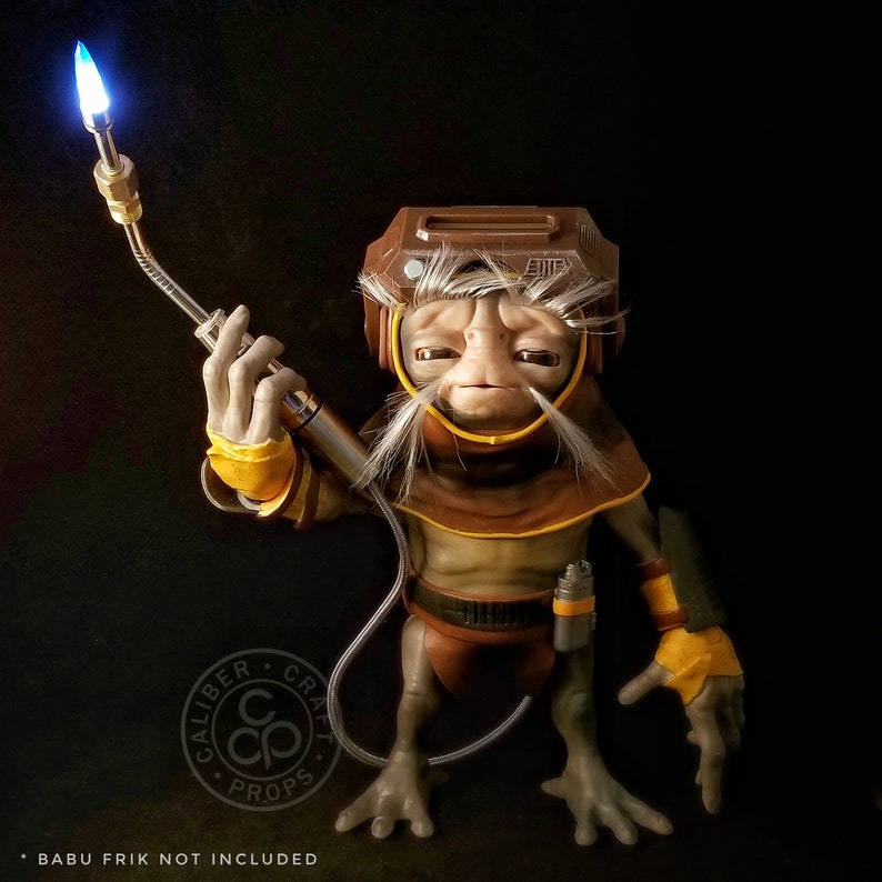 May include: A brown and tan alien figure with a large head and a long, white beard. The figure is holding a silver and black tool in its right hand. The figure is wearing a brown and yellow outfit. The figure is standing on a black background.