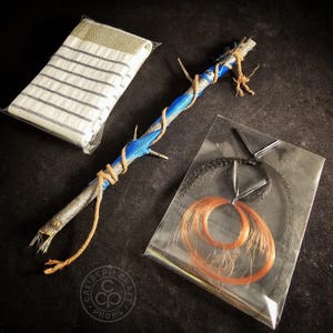 May include: A handmade prop wand with blue ribbon and twine detailing, with a small, striped cloth and a package of orange and black strands. The wand is made from a natural branch.