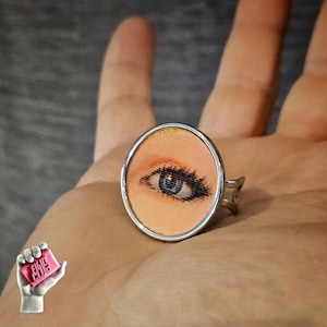 May include: A silver-toned ring featuring a circular setting with a printed graphic of a human eye. The eye has a light peach-colored background, dark eyelashes, and a blue iris. The ring band is thin and metallic with a simple, open design.