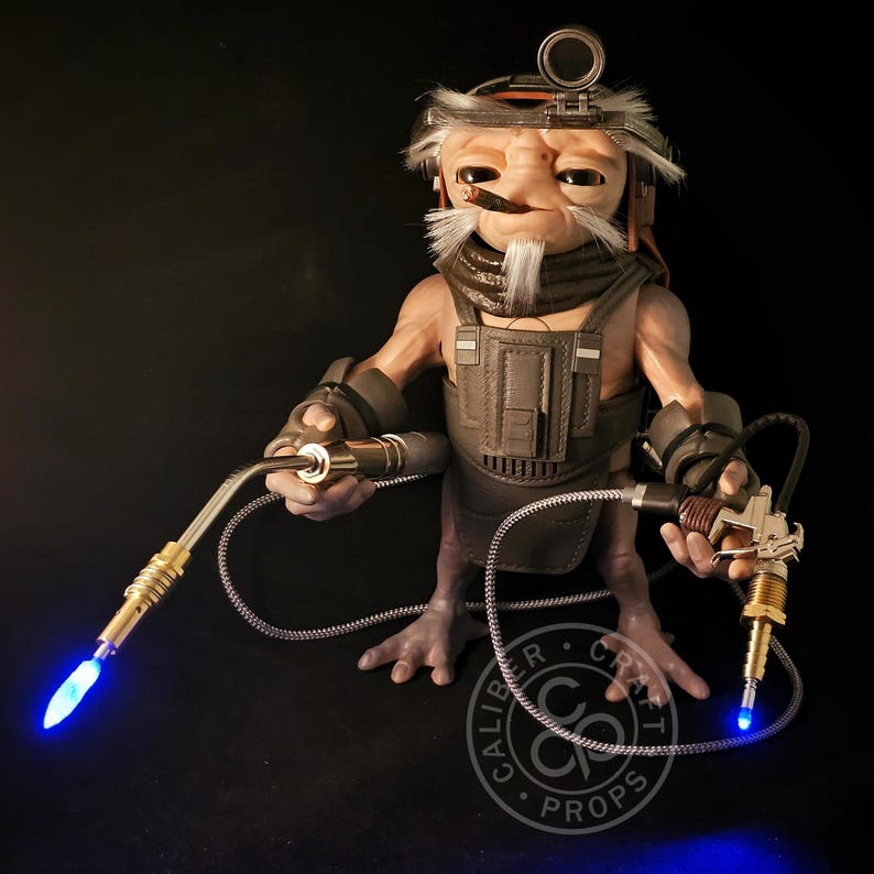 May include: A detailed sculpture of an alien-like figure with a welding torch in each hand. The figure wears a dark vest, goggles, and a white beard. The torches emit a bright blue light, and the sculpture is set against a black background.