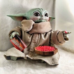 May include: A detailed figurine of a character from a popular science fiction series. The figure is light green with large black eyes, wearing a tan robe. It holds a box of cinnamon candy and a wooden bowl filled with red candies.