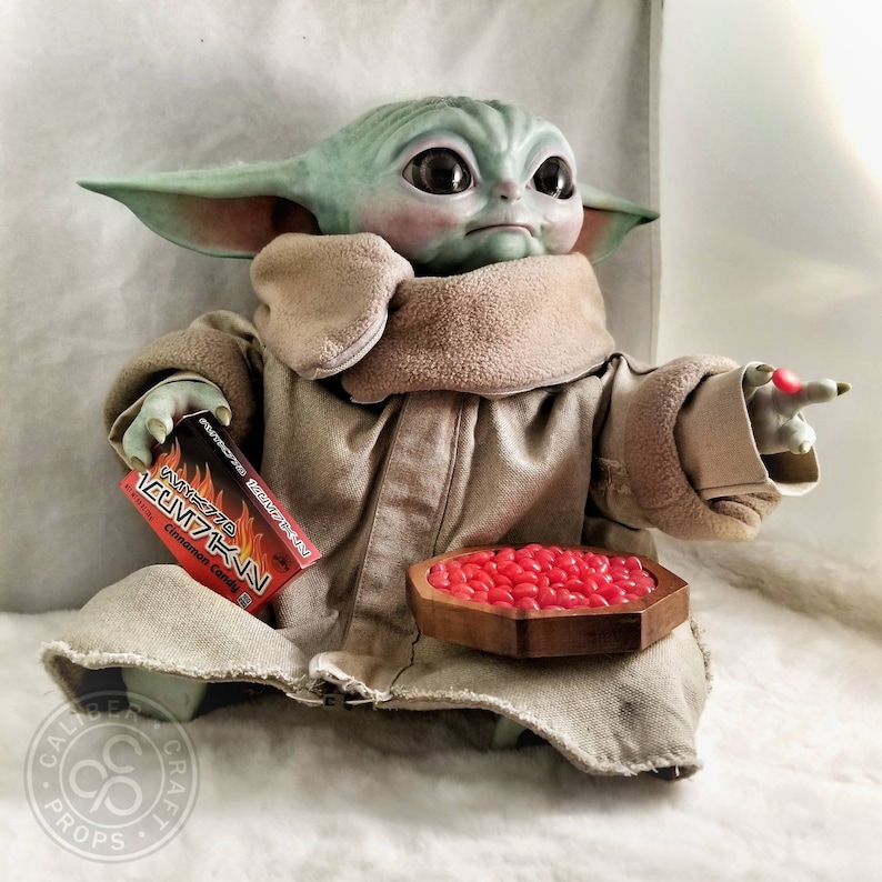 May include: A detailed figurine of a character from a popular science fiction series. The figure is light green with large black eyes, wearing a tan robe. It holds a box of cinnamon candy and a wooden bowl filled with red candies.