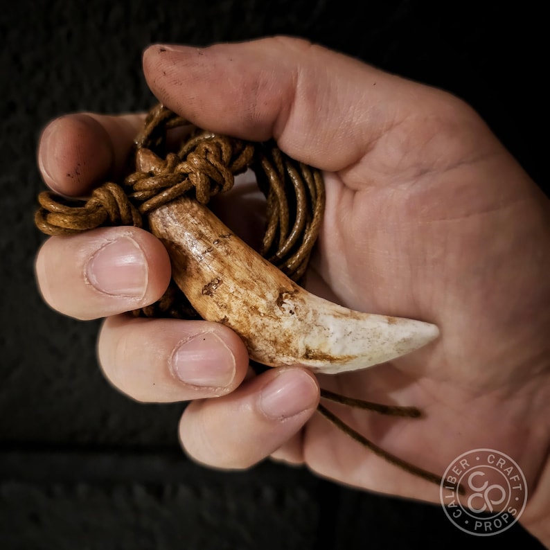 Puede incluir: Un collar con colgante artesanal con un colgante grande, curvado en forma de diente, de color blanquecino. El colgante cuelga de un cord&oacute;n de cuero marr&oacute;n, anudado en la parte superior. El collar tiene una est&eacute;tica r&uacute;stica y natural.
