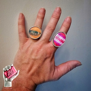 May include: A hand wearing two rings. One ring has a pink and white oval design, the other ring has a round design with a woman's eye.