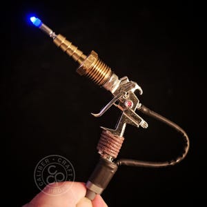 May include: A steampunk-style prop gun with a metallic finish, featuring brass fittings, a silver body, and a blue LED at the barrel's tip. The handle is wrapped with brown twine, and a black tube extends from the base. The logo "Caliber Craft Props" is visible.