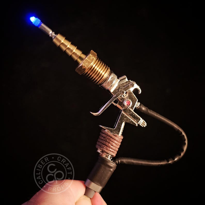 May include: A steampunk-style prop gun with a metallic finish, featuring brass fittings, a silver body, and a blue LED at the barrel's tip. The handle is wrapped with brown twine, and a black tube extends from the base. The logo "Caliber Craft Props" is visible.