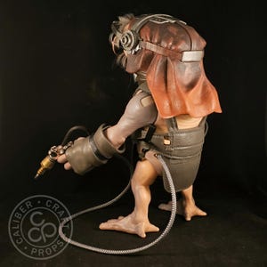 May include: A detailed sculpture of a creature with a brown and red head covering, goggles, and a metallic device in its hand. The figure is wearing a gray harness and gloves, with a cable connecting to the device. The sculpture has a unique, otherworldly appearance.
