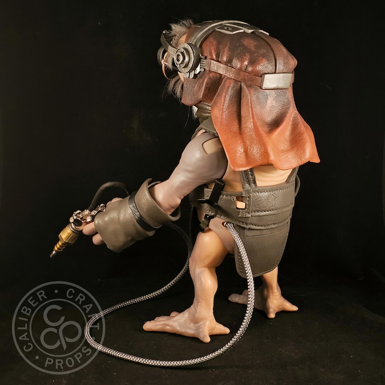 May include: A detailed sculpture of a creature with a brown and red head covering, goggles, and a metallic device in its hand. The figure is wearing a gray harness and gloves, with a cable connecting to the device. The sculpture has a unique, otherworldly appearance.