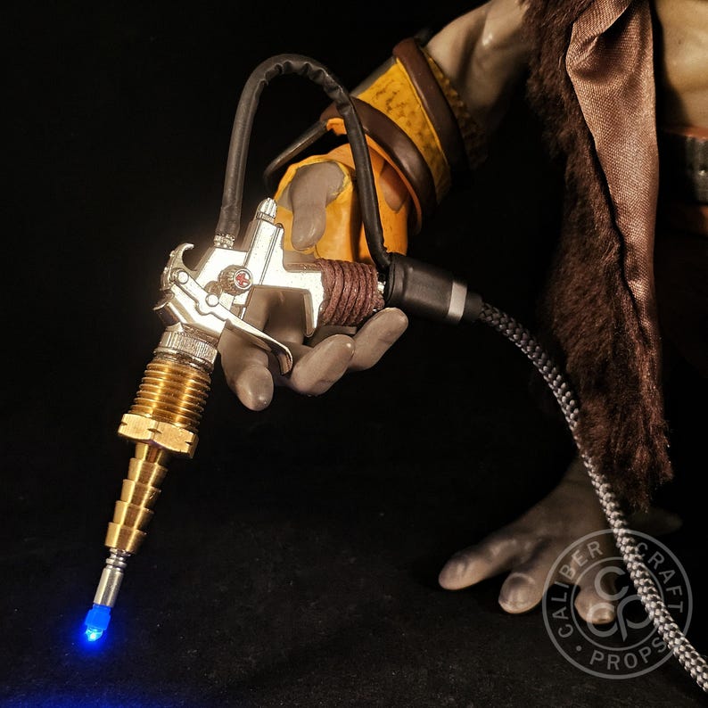 May include: A detailed prop gun with a silver body, brass nozzle, and a blue LED light at the tip. The gun is held by a figure wearing a yellow and brown glove. The prop has a black hose and is labeled with "CALIBER CRAFT PROPS".