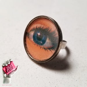 Può includere: Un anello d'argento con un cabochon rotondo in vetro trasparente che presenta un'immagine ravvicinata di un occhio blu.