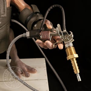 May include: A detailed close-up of a metallic prop gun with a brass nozzle and a brown leather-wrapped handle. The gun is held by a hand, with a black hose and a braided silver cable attached. The background is dark.