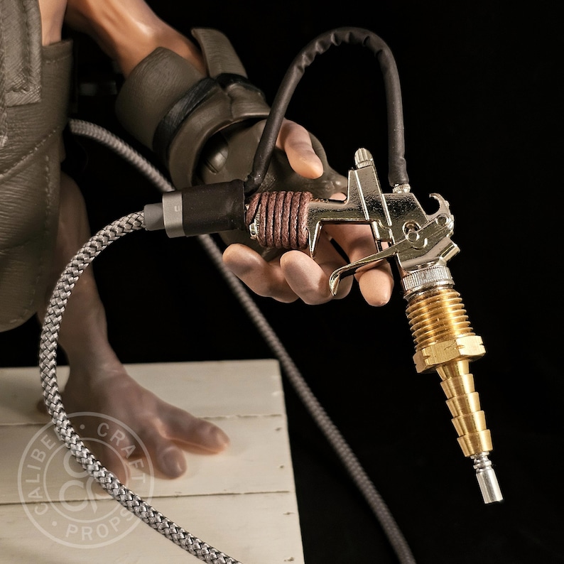 May include: A detailed close-up of a metallic prop gun with a brass nozzle and a brown leather-wrapped handle. The gun is held by a hand, with a black hose and a braided silver cable attached. The background is dark.
