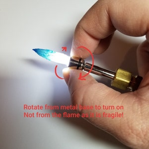 May include: A close-up of a small, handheld torch with a blue flame. The torch has a metal base and a clear glass tube. The text "Rotate from metal base to turn on Not from the flame as it is fragile!" is visible in the image.