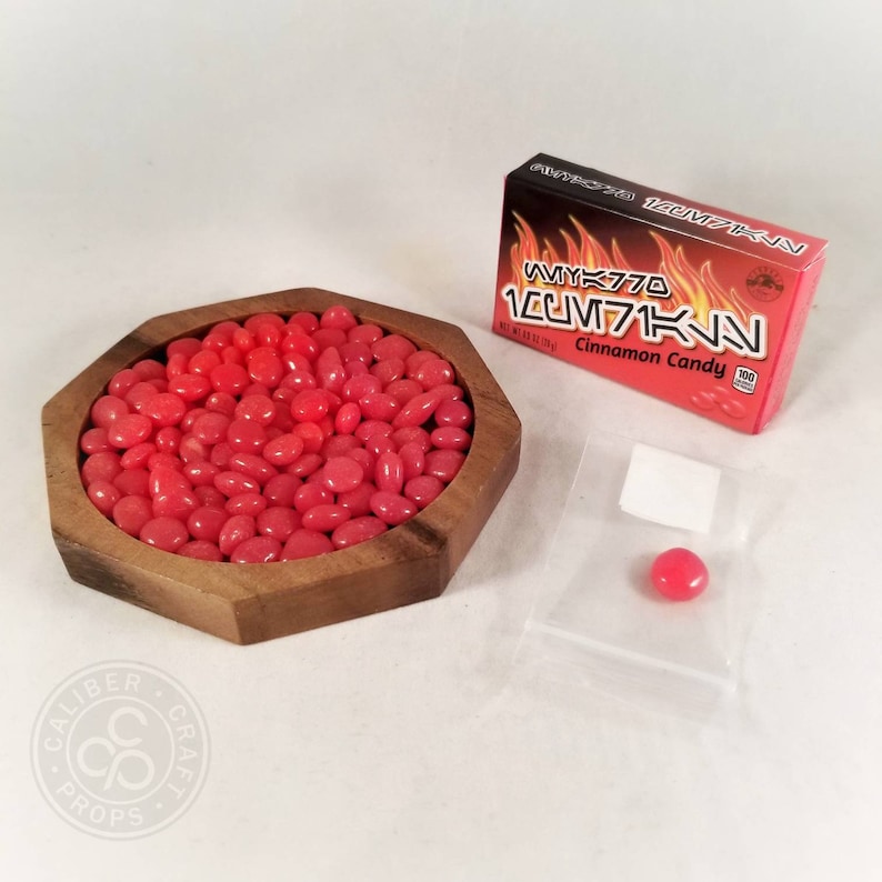 May include: A wooden, octagonal bowl filled with red jelly beans. A box of "WYK770 LUM71KN Cinnamon Candy" is to the right. A single candy is in a small plastic bag.
