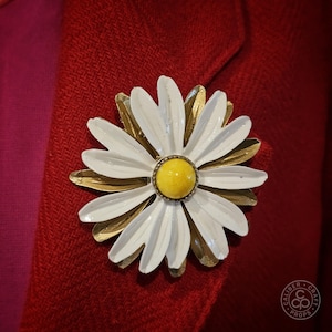 Peut inclure: Grosse broche en forme de marguerite avec des pétales blancs et un centre jaune, sur une base dorée. La broche est épinglée sur un blazer rouge, créant un contraste saisissant. Accessoire vintage.