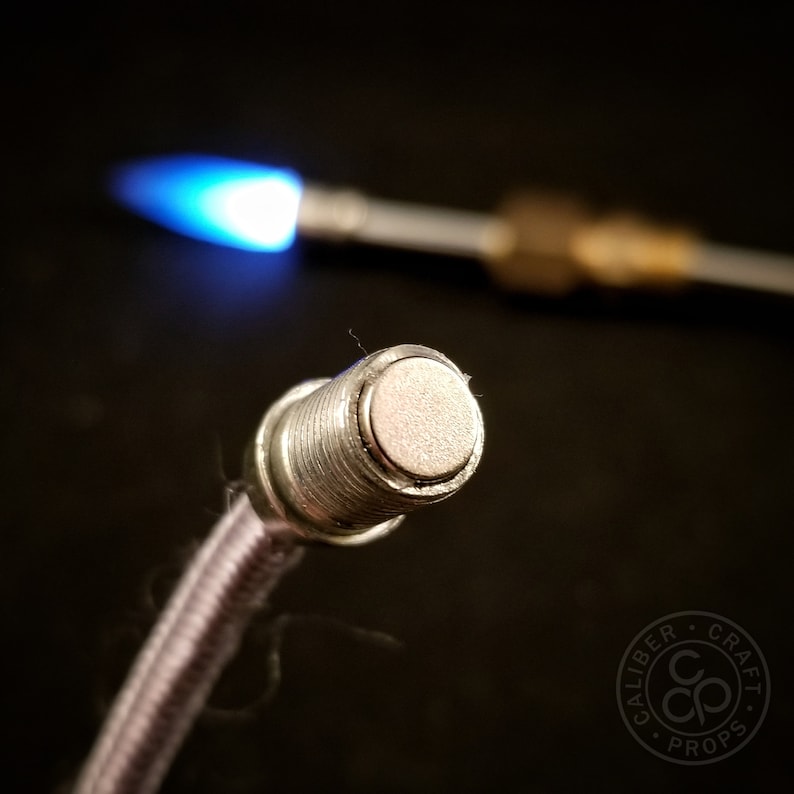 May include: Close-up of a silver metal fitting with a threaded end and a flat, circular top. The fitting is attached to a gray braided hose.