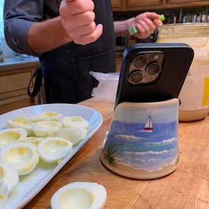 May include: A white ceramic phone stand with a blue and white seascape design. A black iPhone is in the stand. A plate of peeled hard-boiled eggs is on a wooden surface.