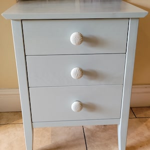 May include: A light blue wooden bedside table with three drawers and white ceramic knobs. The bedside table has a simple design and is perfect for a bedroom.