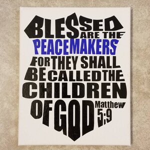 Może przedstawiać: Białe płótno z czarnym i niebieskim wzorem graficznym. Grafika ma kształt tarczy z napisem "Blessed are the Peacemakers for they shall be called the Children of God Matthew 5:9".
