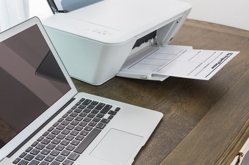 May include: A white printer with a sheet of paper with the text "The Weekly Planner" coming out of the paper tray. A silver laptop computer is on the table to the left of the printer.