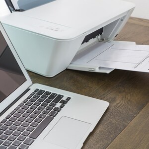 Pode incluir: Uma impressora branca com um documento saindo da bandeja de papel. Um computador port&aacute;til prateado est&aacute; em uma mesa de madeira ao lado da impressora.