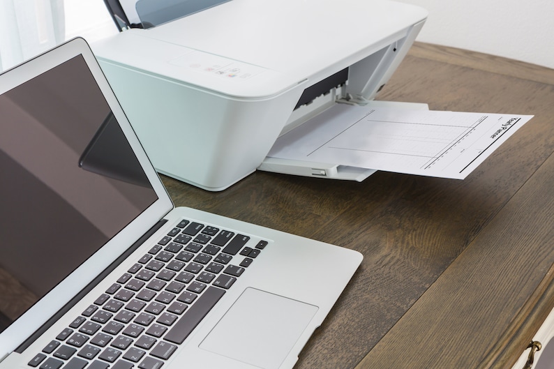 Pode incluir: Uma impressora branca com um documento saindo da bandeja de papel. Um computador port&aacute;til prateado est&aacute; em uma mesa de madeira ao lado da impressora.