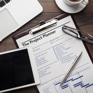 May include: A close-up of a project planner with a brown leather cover and a silver clip. The planner is open to a calendar view with tasks and activities listed. The planner is on a wooden table with a laptop, a cup of coffee, and a pair of glasses.