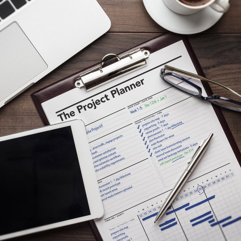 May include: A close-up of a project planner with a brown leather cover and a silver clip. The planner is open to a calendar view with tasks and activities listed. The planner is on a wooden table with a laptop, a cup of coffee, and a pair of glasses.