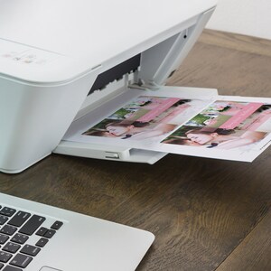 May include: A white printer with a sheet of paper emerging from the paper tray. The paper features a photograph of a couple. The printer is situated on a wooden table alongside a laptop.