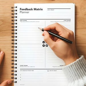 May include: A white spiral-bound Feedback Matrix Planner on a wooden surface. The planner has sections for "Keep Doing," "Stop Doing," "Start Doing," and "Improve." A hand holding a black pen is writing in the planner.