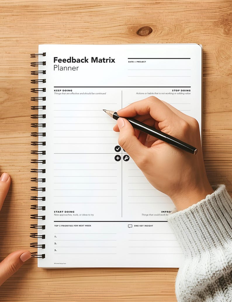 May include: A white spiral-bound Feedback Matrix Planner on a wooden surface. The planner has sections for "Keep Doing," "Stop Doing," "Start Doing," and "Improve." A hand holding a black pen is writing in the planner.