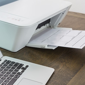 Peut inclure: Une imprimante blanche avec un bac &agrave; papier &eacute;tendu, imprimant une feuille de papier avec un design de planificateur. Un ordinateur portable est sur la table &agrave; gauche de l'imprimante.