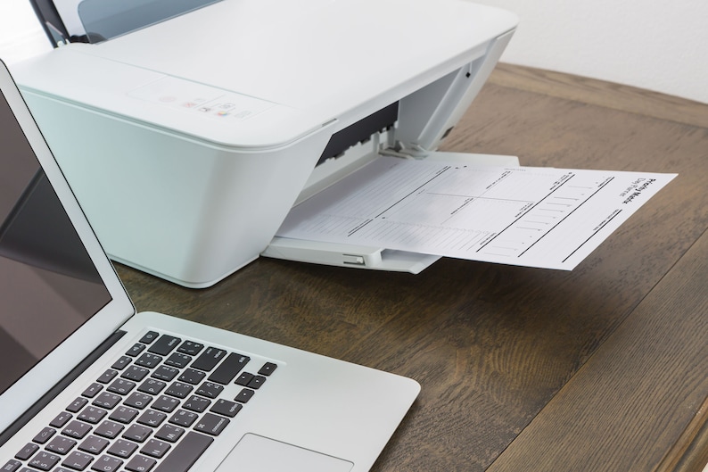Peut inclure: Une imprimante blanche avec un bac &agrave; papier &eacute;tendu, imprimant une feuille de papier avec un design de planificateur. Un ordinateur portable est sur la table &agrave; gauche de l'imprimante.