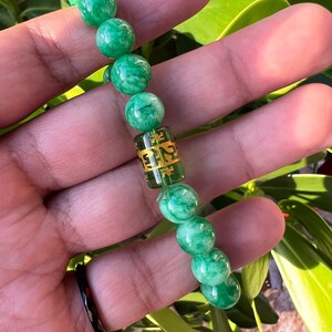 May include: Green jade bead bracelet with a gold charm featuring Chinese characters. The bracelet is on a hand.
