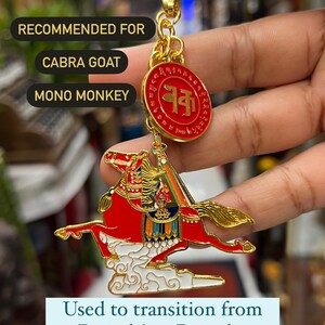 Pode incluir: Um amuleto de cavalo de vento dourado com detalhes vermelhos e brancos. O amuleto está preso a uma corrente dourada e apresenta um pingente redondo com um círculo vermelho e caracteres chineses. O amuleto é recomendado para a cabra Cabra e o macaco Mono.