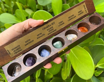 May include: A wooden box with seven compartments containing different coloured stones. The box is labelled with the names of the seven chakras and the corresponding stones. The stones are clear quartz, amethyst, sodalite, green aventurine, citrine, carnelian, and red garnet.