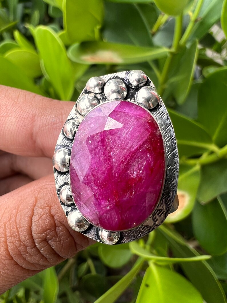 May include: A silver ring featuring a large, faceted, pink gemstone set in a textured bezel. The ring is surrounded by small, round silver beads.