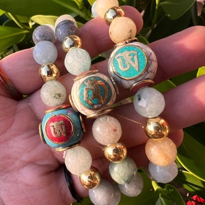 May include: Three bracelets with gold beads and charms. The charms are round and feature a turquoise and gold design. The bracelets are made of different colored stones, including light blue, gray, and beige.