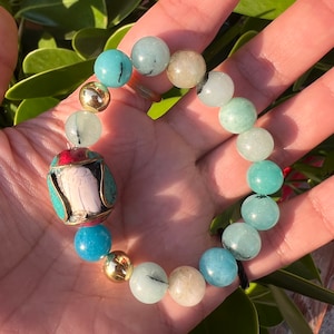 May include: A turquoise, green, and white beaded bracelet with a gold accent and a decorative charm featuring a white figure with red and turquoise accents.