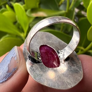 May include: A silver ring featuring a large, oval-shaped, deep pink gemstone set in the centre. The ring is crafted from hammered silver and has a textured finish.
